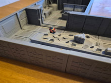 Load image into Gallery viewer, A miniature tabletop gaming terrain piece depicting a sci-fi corridor or room, shown on a wooden surface. The gray plastic structure features a small figure of a character in a red and blue outfit standing on a platform.