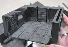 Load image into Gallery viewer, A miniature 3D-printed room terrain piece for tabletop wargaming, held by a hand. The model features detailed paneling, a tiled floor, and a dark gray color, shown against a blurred background of other miniature pieces.