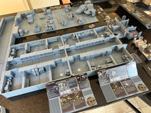 Load image into Gallery viewer, A tabletop display of a miniature model terrain set, likely for tabletop wargaming. The main subject is a large, multi-level gray modular base or city map with detailed room layouts, accompanied by two printed character or unit cards in the foreground and other gaming accessories on a dark reflective surface.